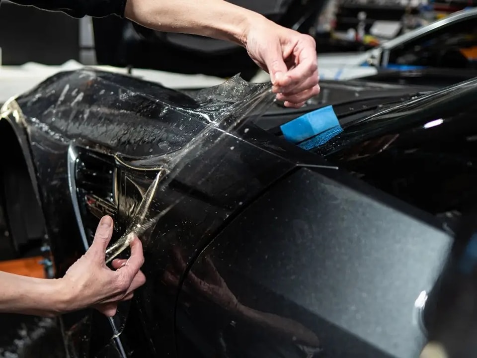 "Close-up of a person applying Paint Protection Film (PPF) to a car's surface, focusing on the smooth, clear film being carefully positioned and pressed against the car's paint.